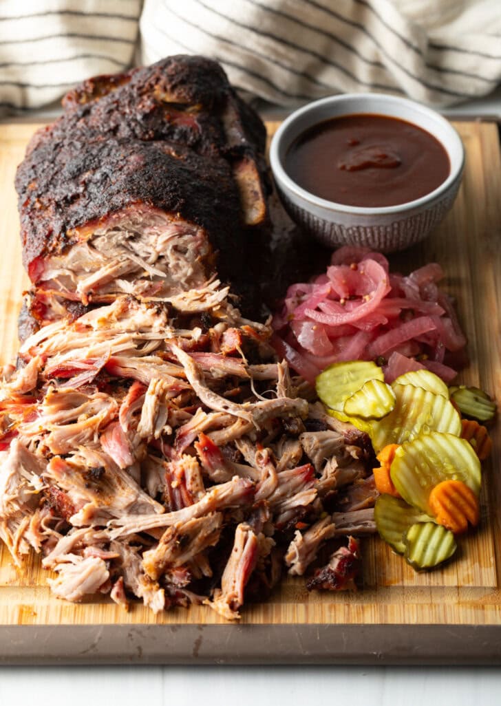 Shredded smoked pork butt on a cutting board, with ramekin of bbq sauce, stack of pickles, and pickled red onions.