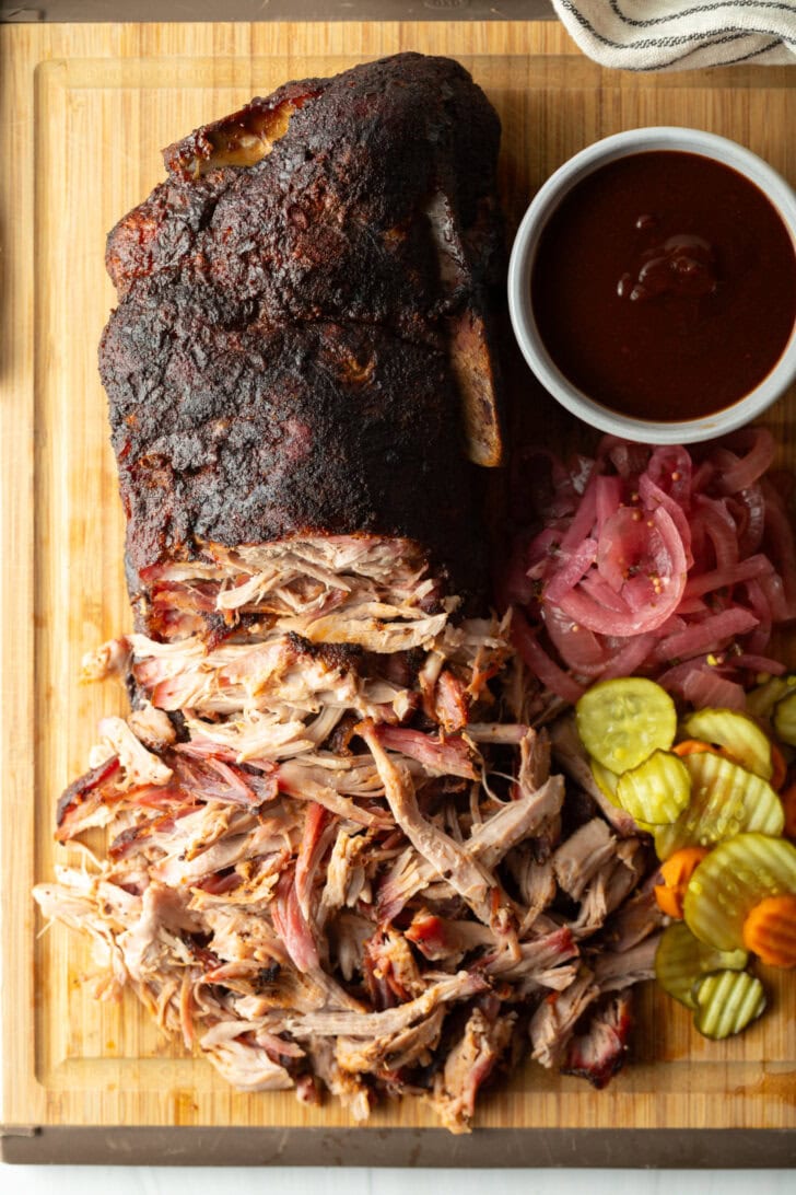 Shredded smoked pork butt on a cutting board, with ramekin of bbq sauce, stack of pickles, and pickled red onions.