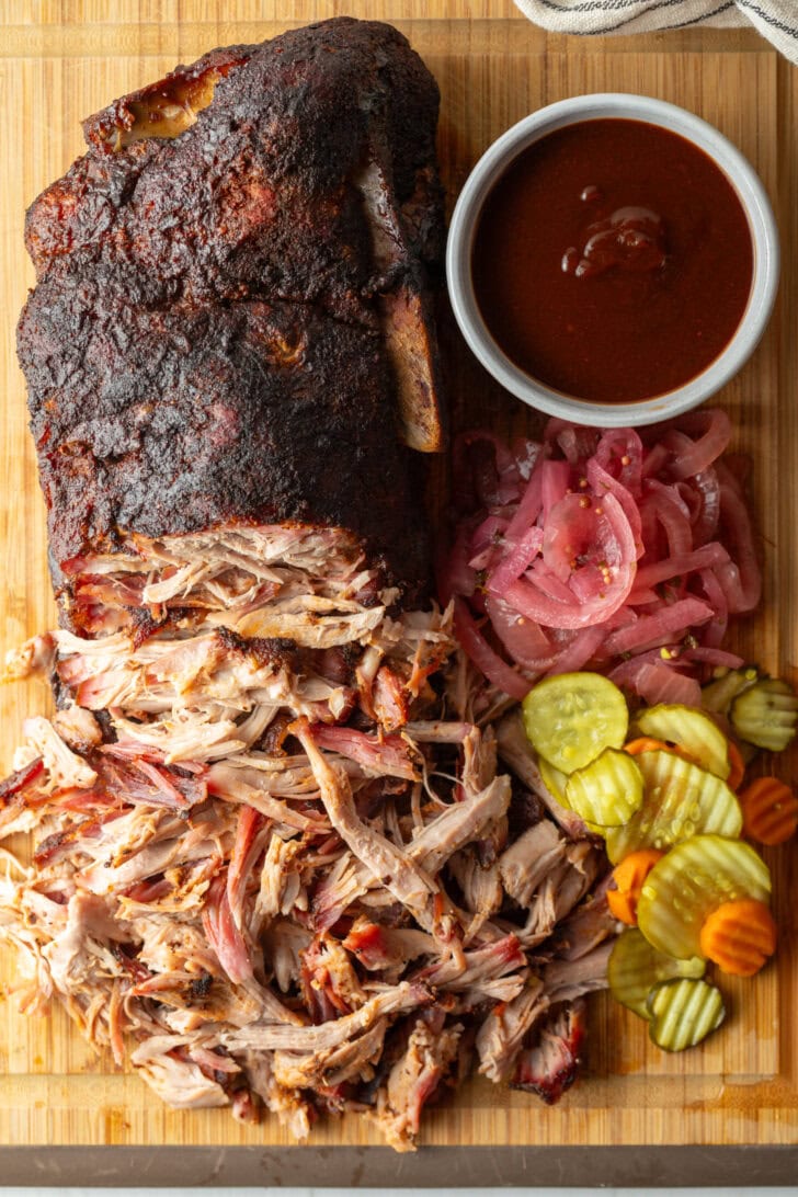 Shredded smoked pork butt on a cutting board, with ramekin of bbq sauce, stack of pickles, and pickled red onions.