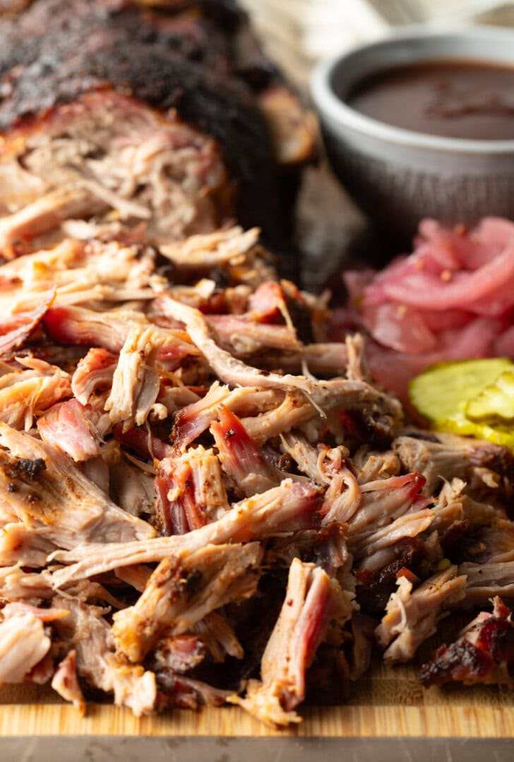 Shredded smoked pork butt on a cutting board, with ramekin of bbq sauce, stack of pickles, and pickled red onions.