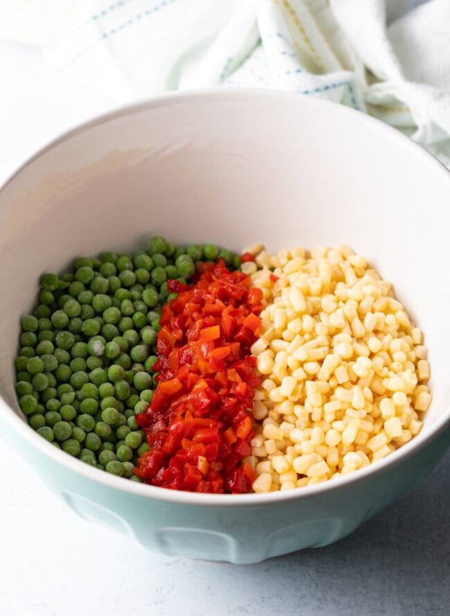 Frozen corn and canned peas, chopped pimentos in a white bowl.