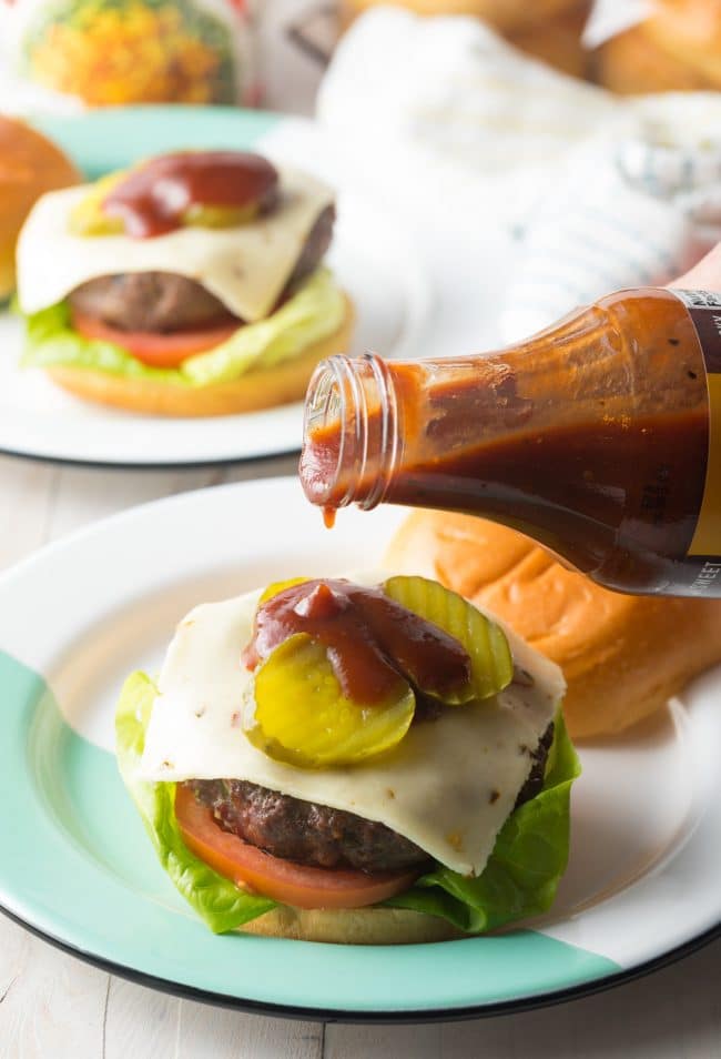 Hand holding a bottle of BBQ sauce and pouring it onto a cowboy burger. 