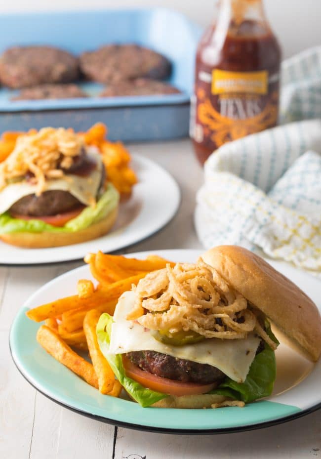 Open faced spicy burger with fried onions on top, next to a pile of french fries. 