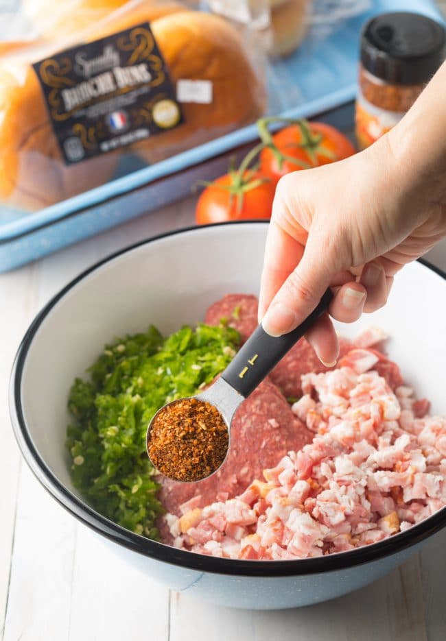 Hand holding a measuring spoon with spices in it over the mixture of raw beef. 