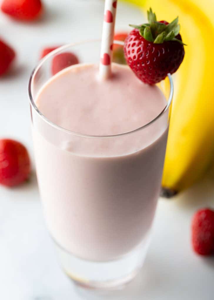 Tall glass of strawberry and banana smoothie, garnished with a whole strawberry on the rim and a red and white straw.