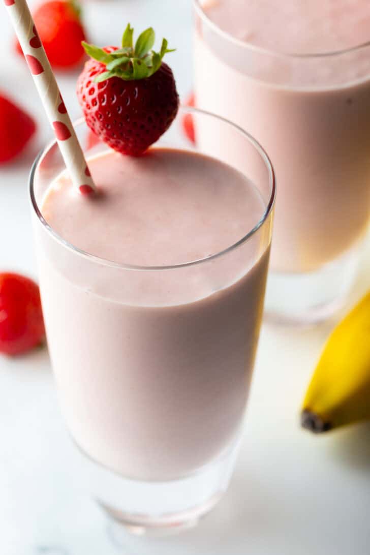 Two tall glasses of strawberry banana smoothies, each garnished with a whole strawberry on the rim and a red and white straw.