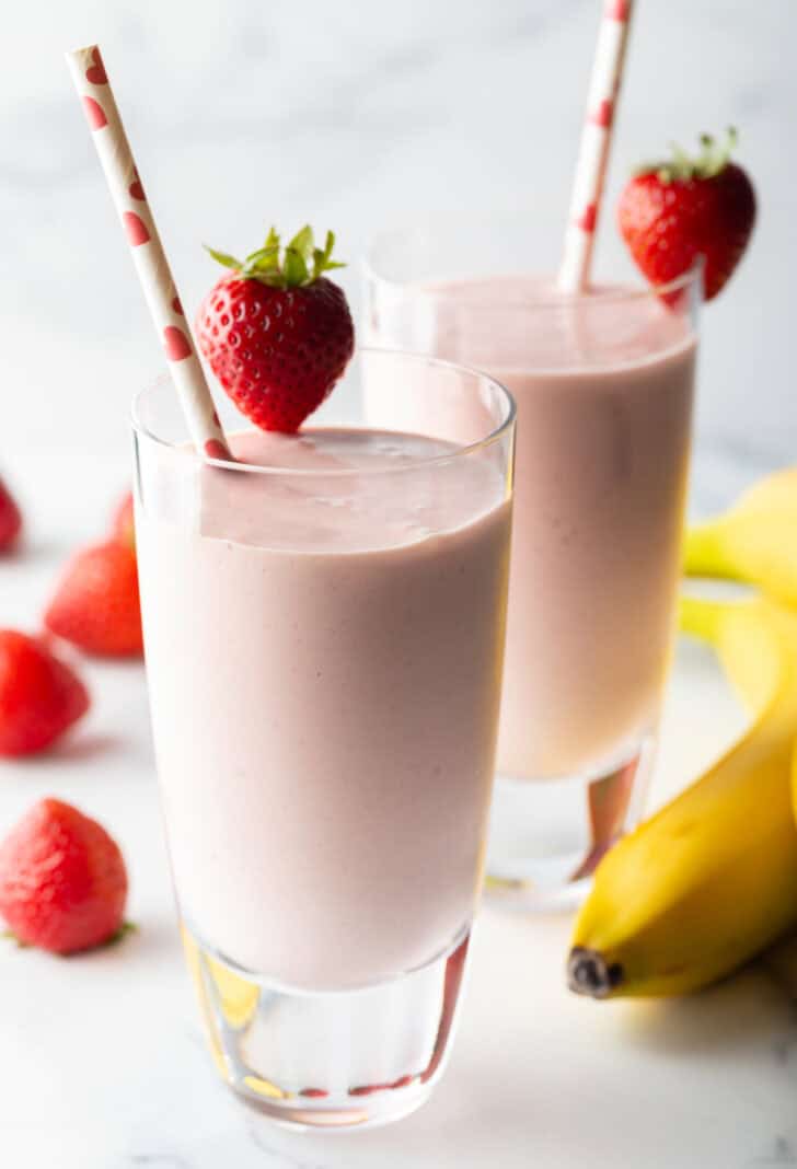 Two tall glasses of strawberry banana smoothies, each garnished with a whole strawberry on the rim and a red and white straw.