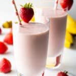 Two tall glasses of strawberry banana smoothies, each garnished with a whole strawberry on the rim and a red and white straw.