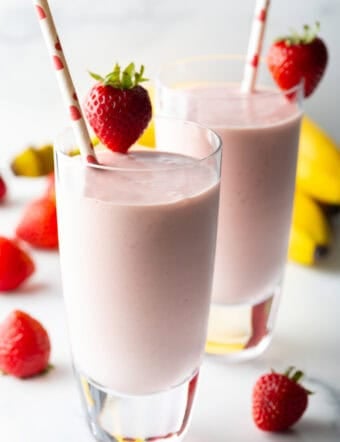 Two tall glasses of strawberry banana smoothies, each garnished with a whole strawberry on the rim and a red and white straw.