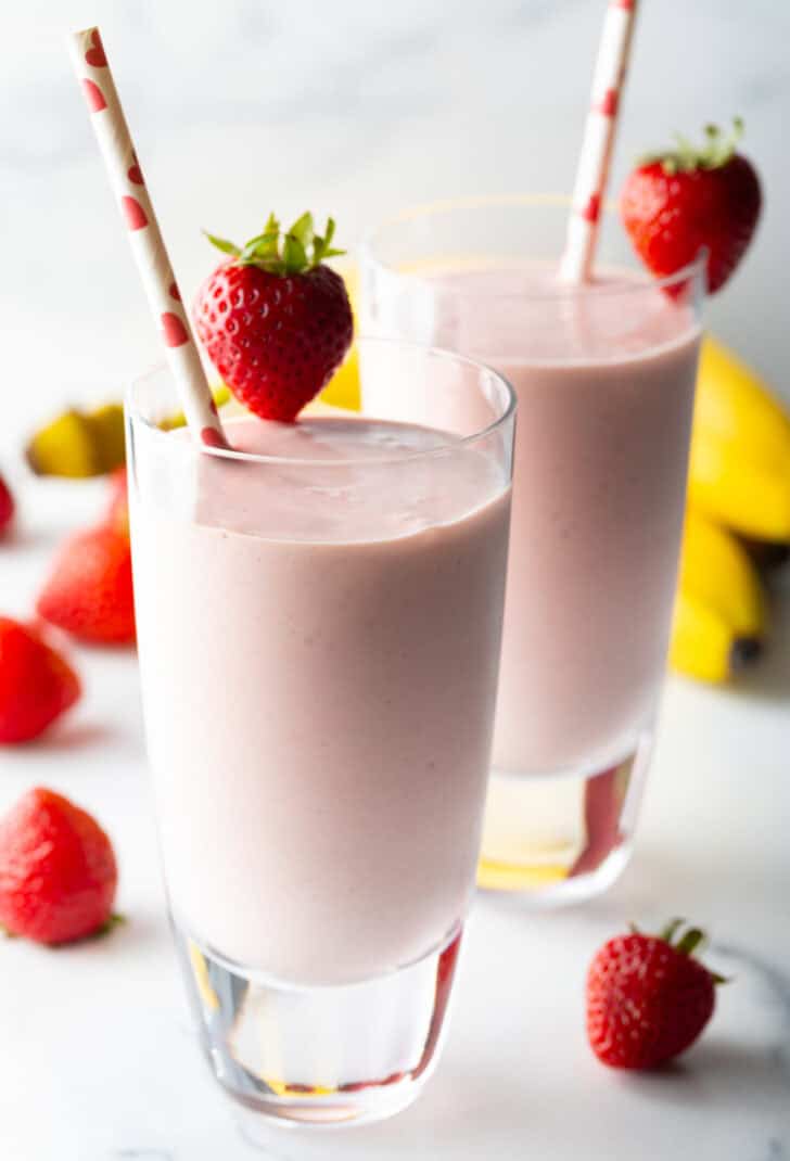 Two tall glasses of strawberry banana smoothies, each garnished with a whole strawberry on the rim and a red and white straw.