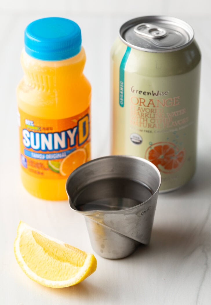 Recipe ingredients: Bottle of juice, metal cup with vodka, fresh orange wedge, and can of soda.