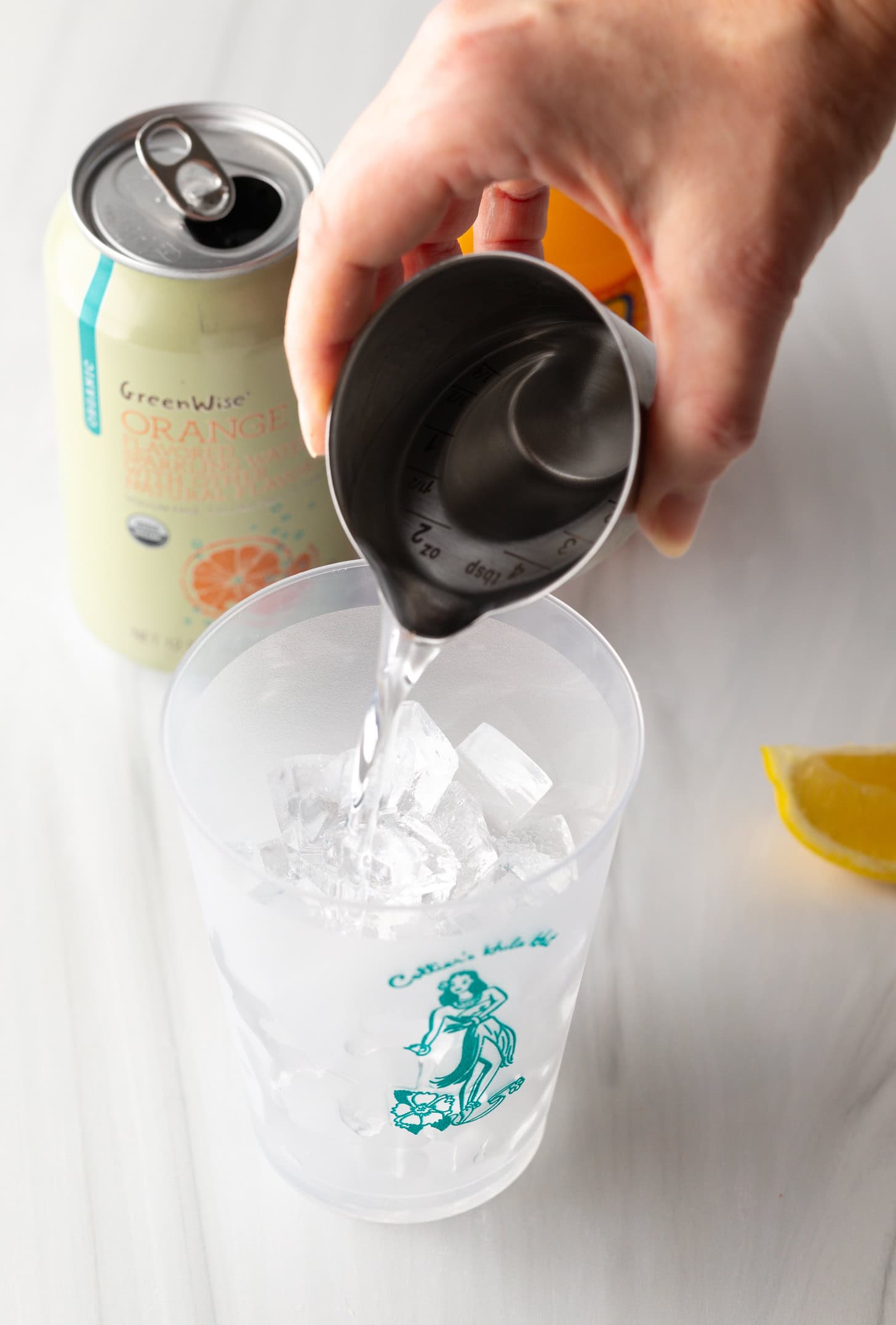 Hand pouring vodka from a metal shot glass into a glass filled with ice.