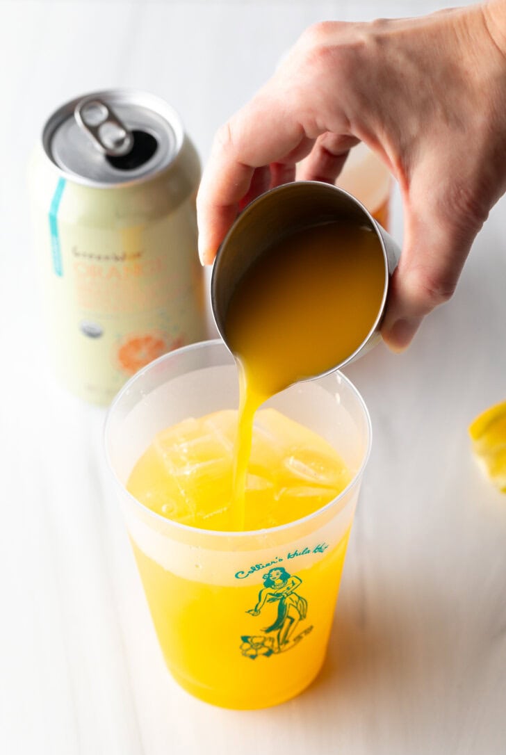 Hand adding orange juice from a metal shot glass into a glass cup.