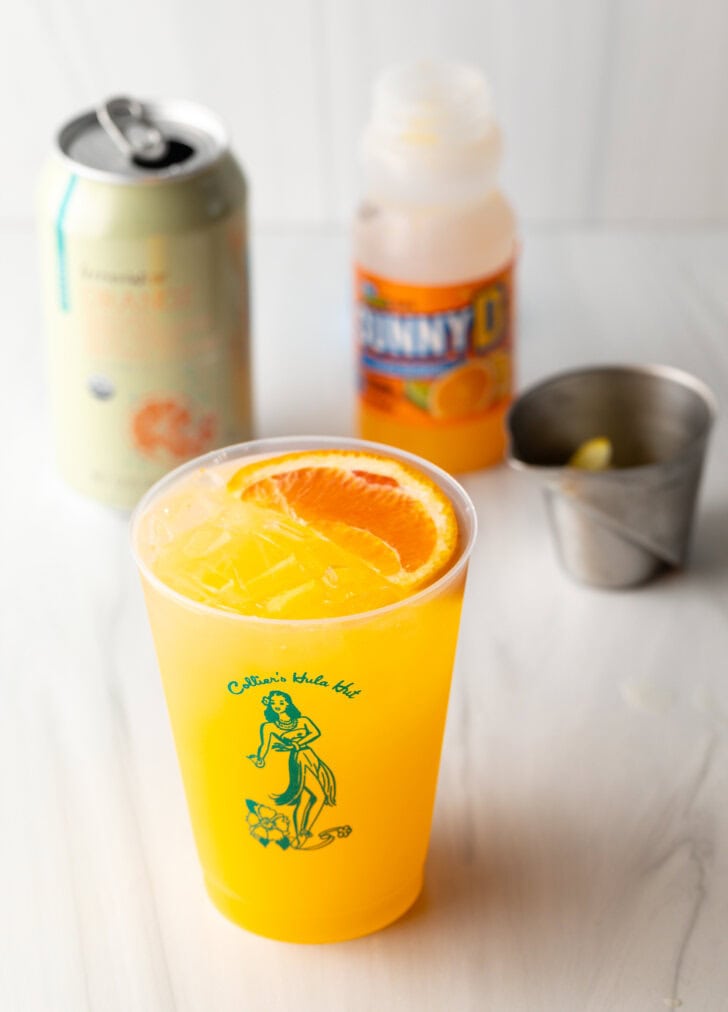 Sunny d vodka seltzer drink in a glass cup with slice of orange on top. Bottle of juice, metal cup with vodka, and can of soda in the background.