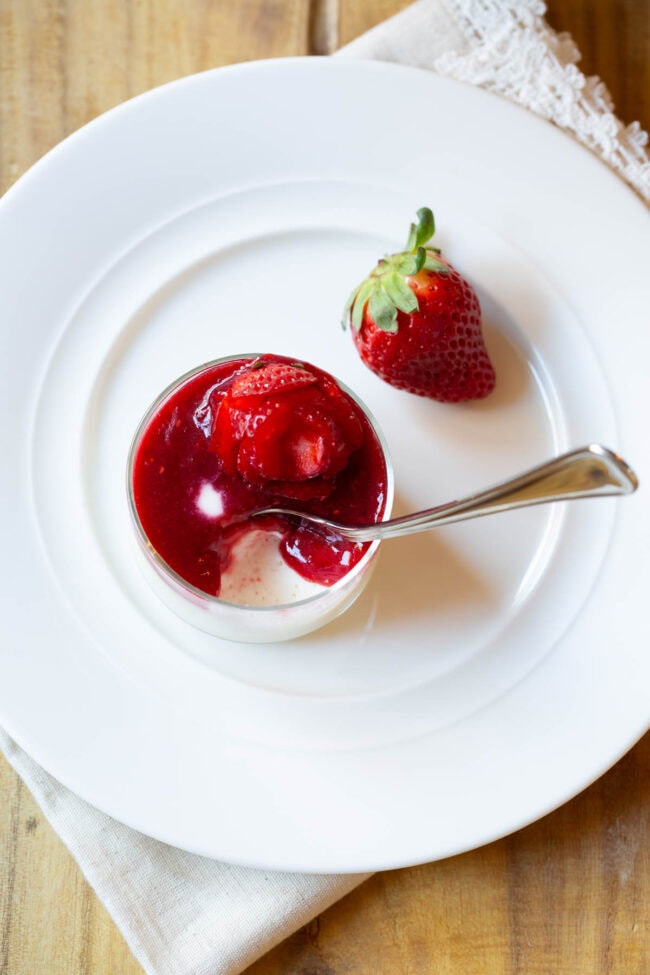 Top down view small glass dish with silky swedish creme topped with berry compote and strawberry rose. A metal spoon is scooping a spoonful from the dish.