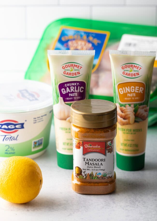 Recipe ingredients on a white background: Containers of yogurt, garlic paste, ginger paste, masala seasoning, chicken, and a fresh whole lemon.