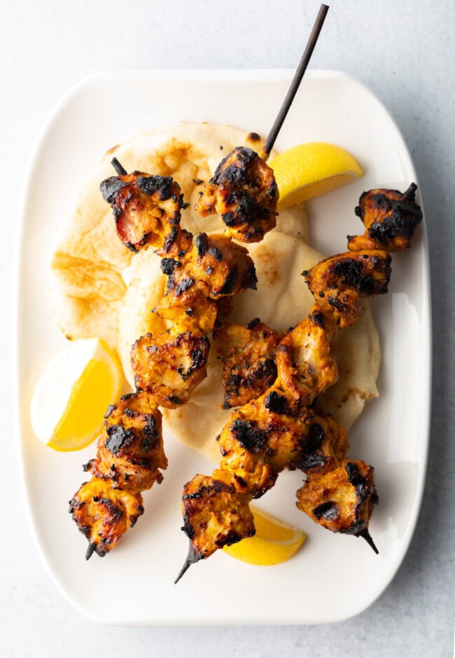 Three tikka chicken kebabs on a white plate with lemon wedges.