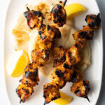 Three tikka chicken kebabs on a white plate with lemon wedges.