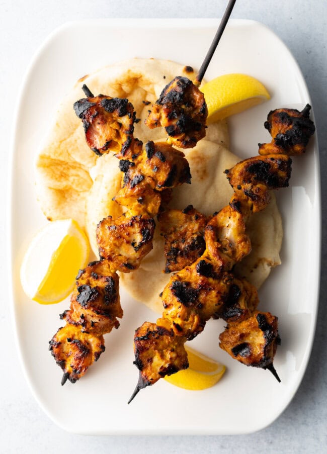 Three tikka chicken kebabs on a white plate with lemon wedges.