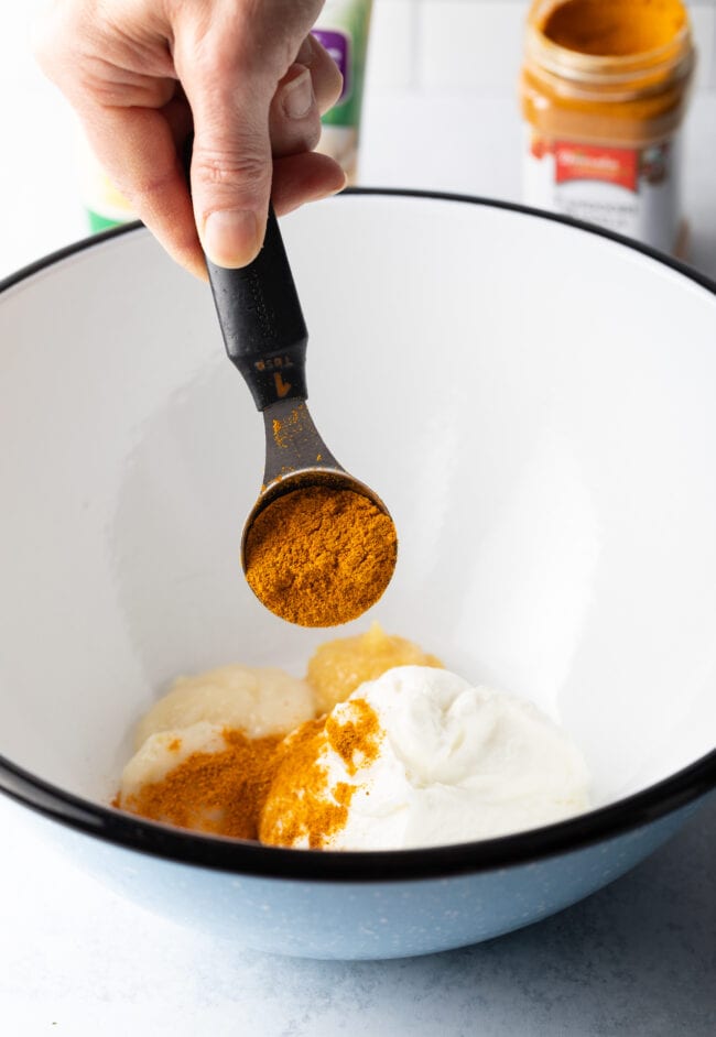 Hand with metal measuring spoon pouring spices into a white bowl with yogurt.