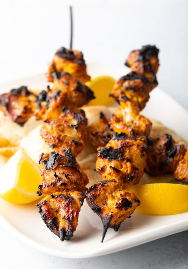 Two tandoori chicken tikka skewers being stood on their ends on a white plate with lemon wedges.