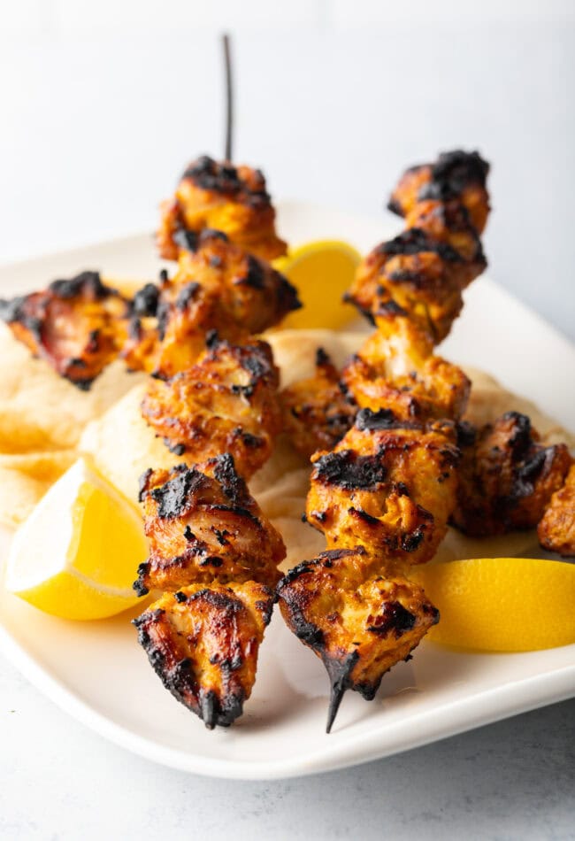 Two tandoori chicken tikka skewers being stood on their ends on a white plate with lemon wedges.