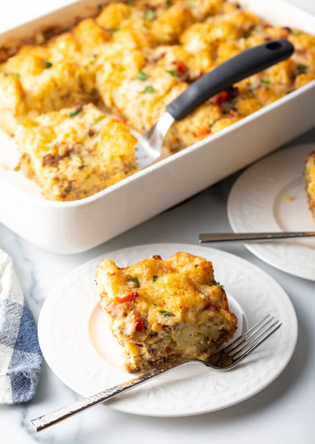 Slice of tater tot casserole on a white plate, full casserole in the background.