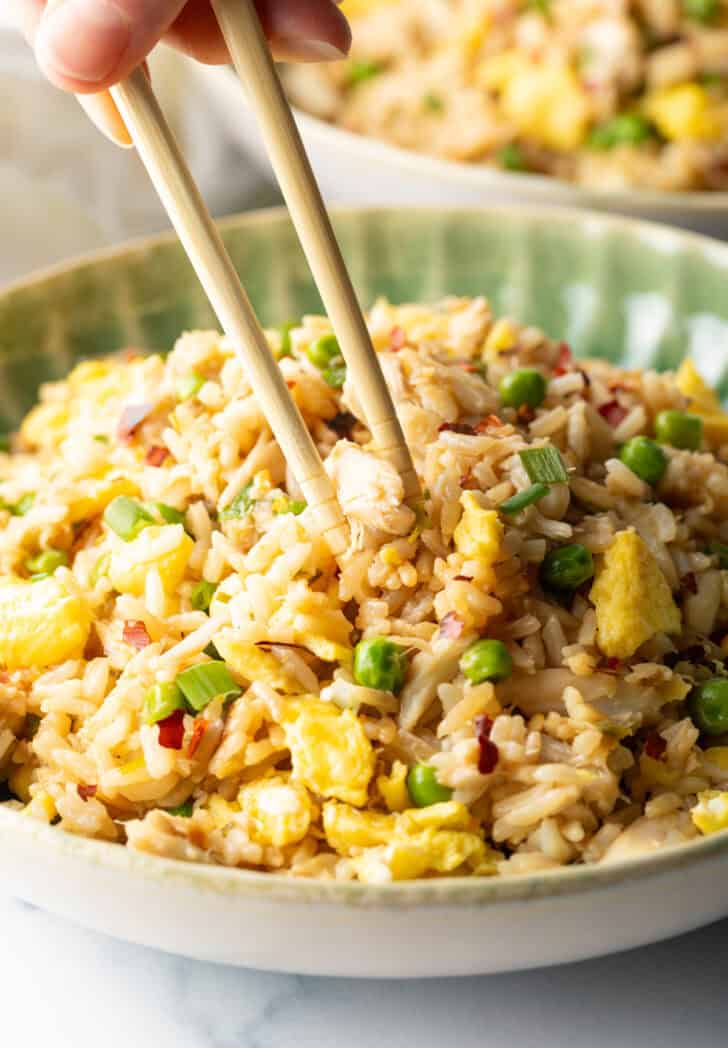 Chopsticks holding a bite of fried rice with crab, taking it from a plate of the rice.