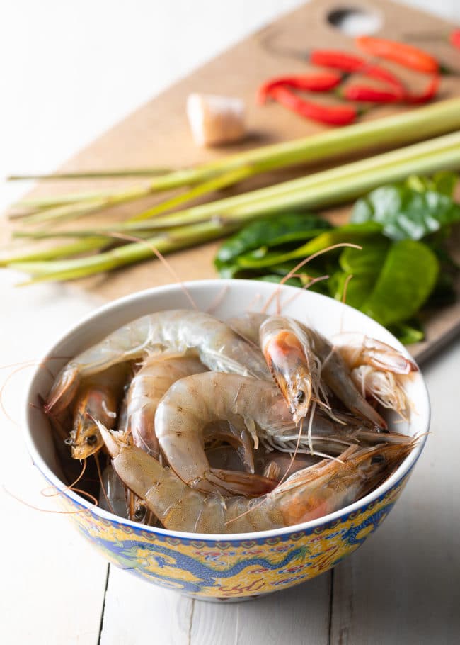 Bowl of raw shrimp with other ingredients for soup in the background. 