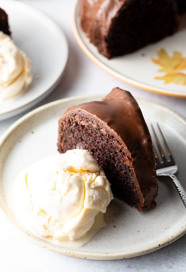 Slice of chocolate glazed cake on a white plate, with serving of vanilla ice cream and fork on the plate.