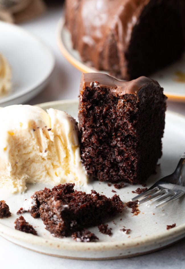 Slice of chocolate glazed cake on a white plate, with serving of vanilla ice cream and fork on the plate.