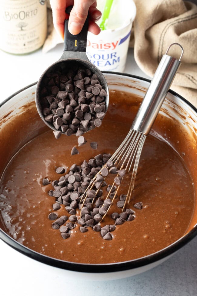 Adding chocolate chunks from a metal measuring scoop to the glass mixing bowl with batter.