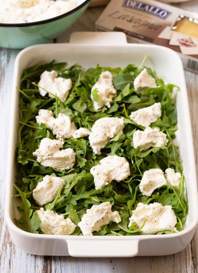 White baking dish with a layer or arugula and dollops of ricotta on top. 