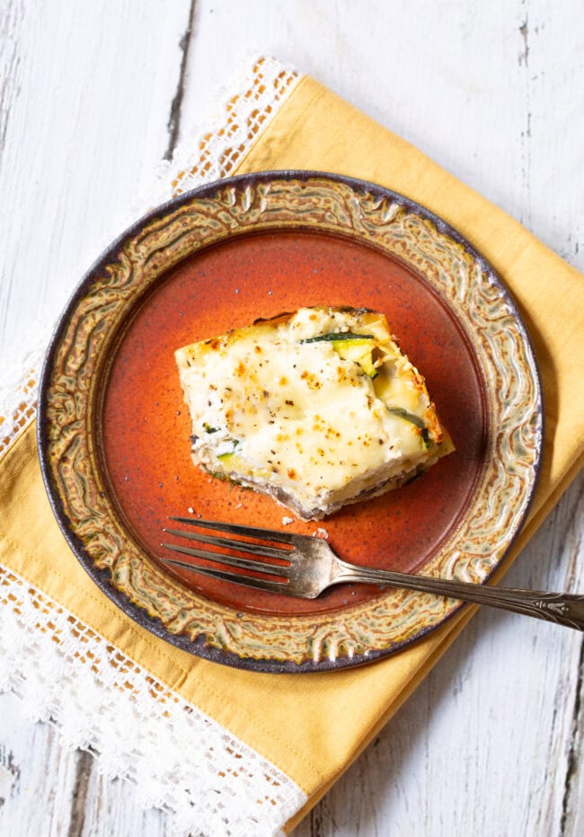 Slice of vegetable lasagna with white sauce on a red plate with a bite missing. 