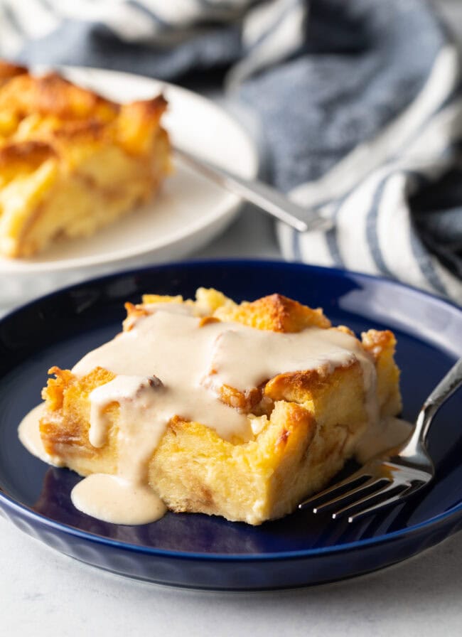 Square of white chocolate bread pudding with vanilla sauce on a blue plate with fork on the side.