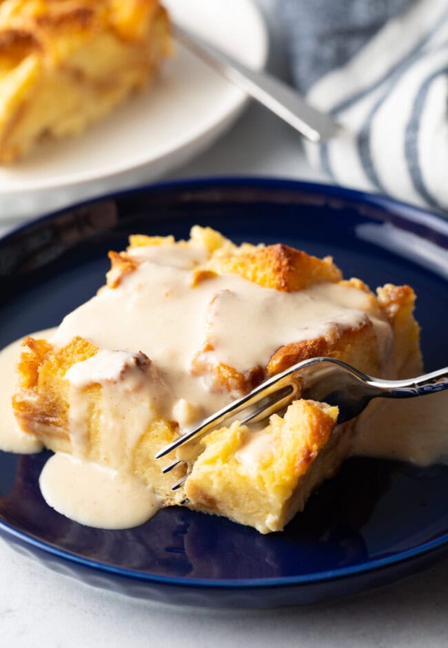 Fork taking a bite from the homemade white chocolate brioche bread pudding with decadent vanilla sauce.