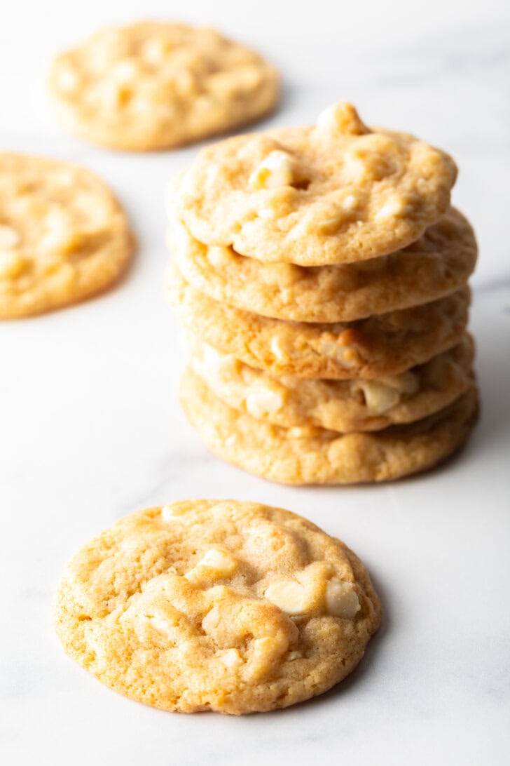 A stack of 5 macadamia nut cookies with white chocolate chips. Another cookie is laying in front of the stack.